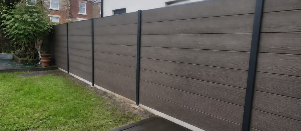 Grey-brown composite boards slotted into slim aluminium posts along a small garden, seen from the edge of a damp composite deck