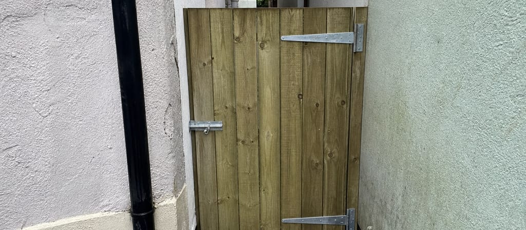 A new closeboard side gate hung on galvanised tee hinges in a rendered alley opening, squared to the gap above a wet path