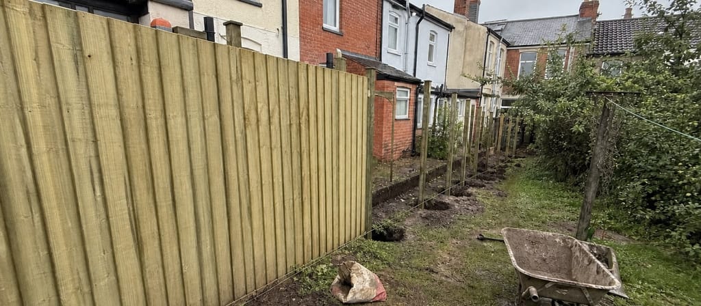 Half a new closeboard run standing pale and fresh in a narrow terrace garden, the remaining post holes dug along a taut string line with a muddy wheelbarrow on the grass