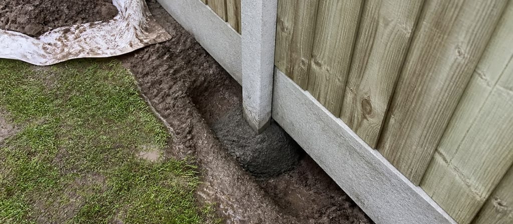 A concrete post and concrete gravel board set at the foot of a closeboard run, the narrow trench alongside still open and spoil heaped on a sheet